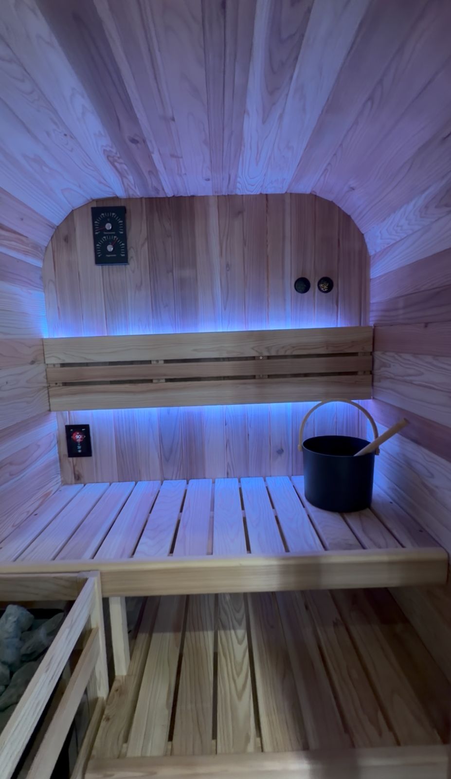 Interior view of a modern sauna with light wood construction, featuring two tiered benches, blue ambient lighting along the walls and ceiling, control panels with dials and buttons, and a black water bucket with wooden ladle on the upper bench.