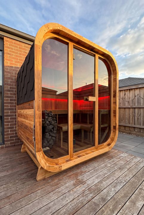 A modern wooden sauna pod with curved glass doors and red interior lighting, positioned on a wooden deck next to a brick building and wooden fence.