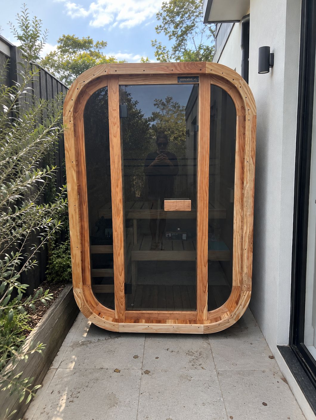 A wooden pod structure with rounded corners and dark tinted glass panels, positioned on a concrete patio between a white building and garden fence on a sunny day.