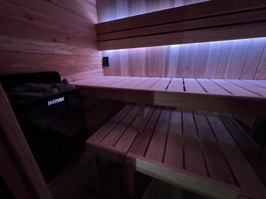 Interior of a modern sauna with wooden benches on two levels, ambient blue and purple LED lighting, and a Harvia sauna heater visible on the left side.