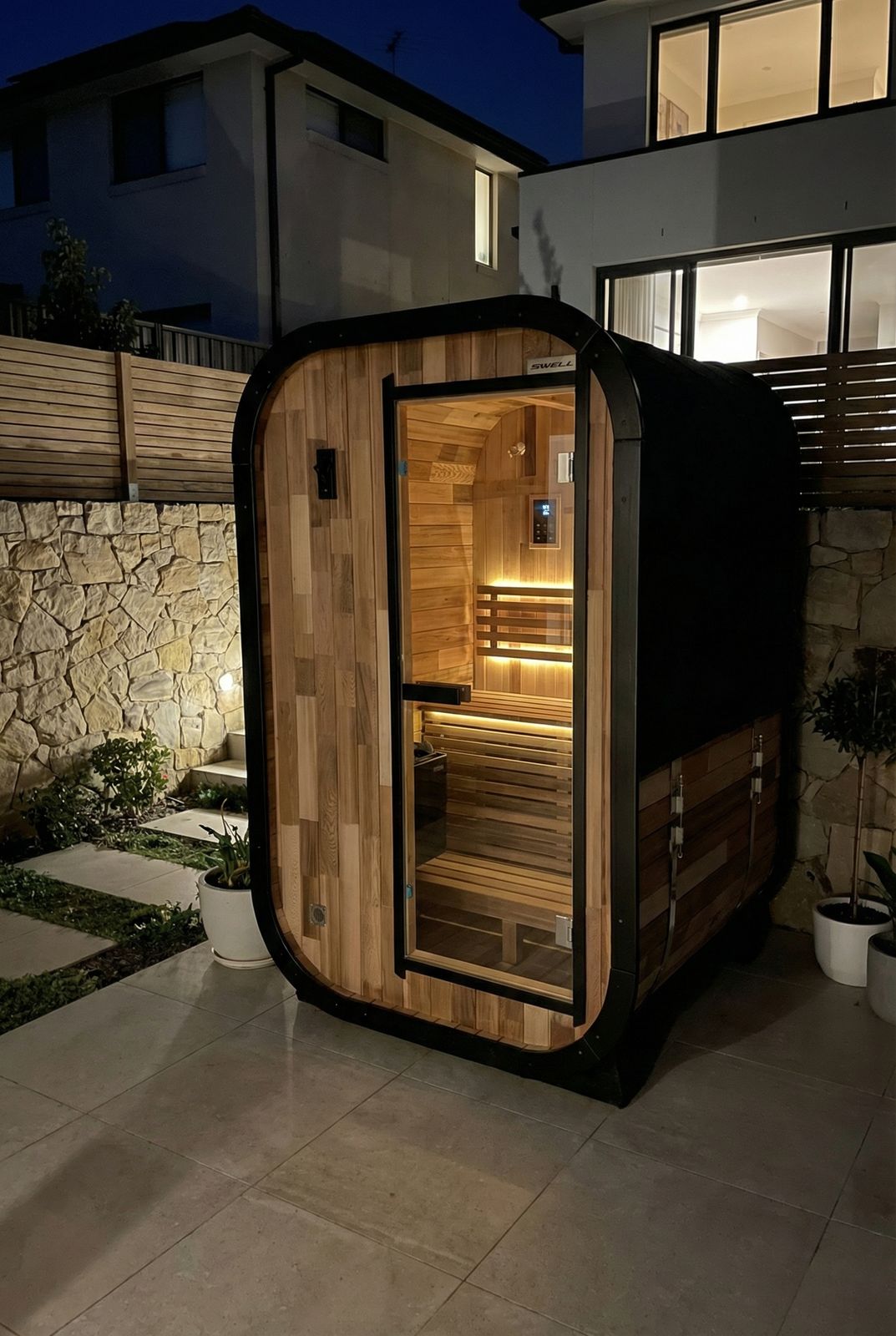 Modern outdoor sauna with curved black exterior and wood interior, illuminated at night in a residential backyard setting