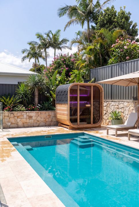 A modern backyard with a rectangular swimming pool, wooden sauna pod with purple lighting, lounge chairs, tropical plants including palm trees and flowering shrubs, stone walls, and contemporary architecture with metal fencing.