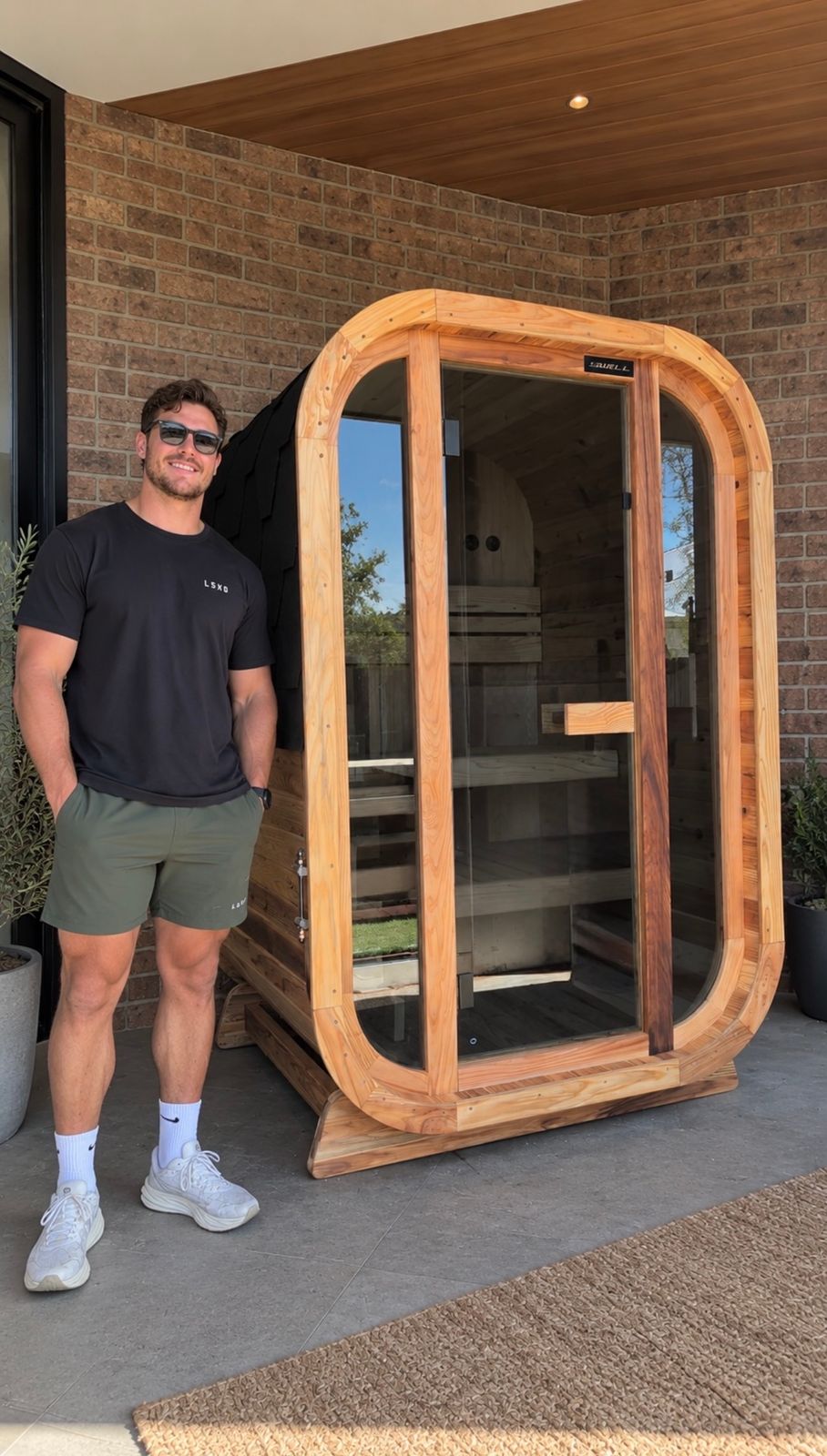 A man wearing sunglasses and casual clothing stands next to a wooden sauna pod with curved glass doors on a patio against a brick wall.