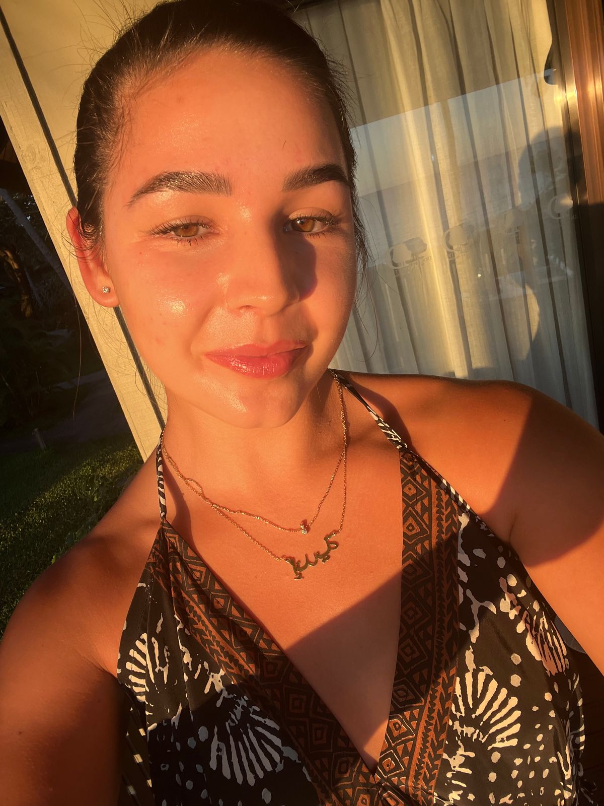 A woman taking a selfie outdoors in golden sunlight, wearing a patterned halter top and layered necklaces, with curtains visible in the background.