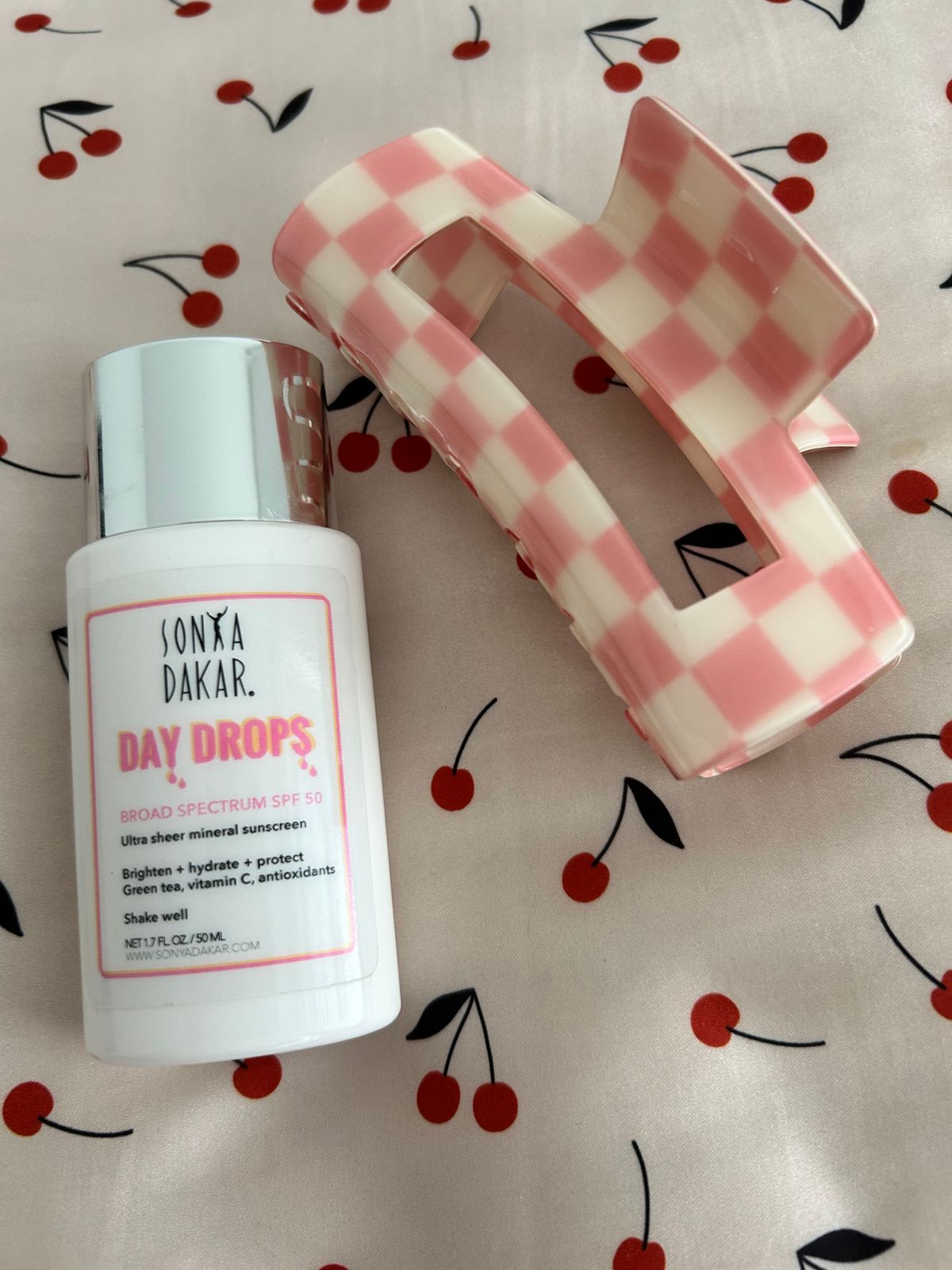 A bottle of Sonya Dakar Day Drops sunscreen with SPF 50 sits next to pink and white checkered hair clips on a white surface decorated with red cherry patterns.