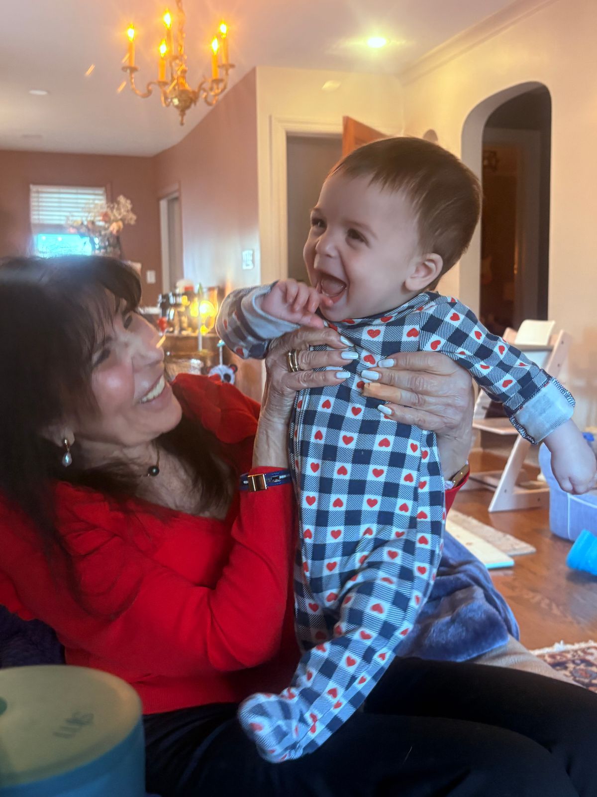 A smiling toddler in heart-patterned pajamas being held by an adult in a red sweater in a warmly lit living room