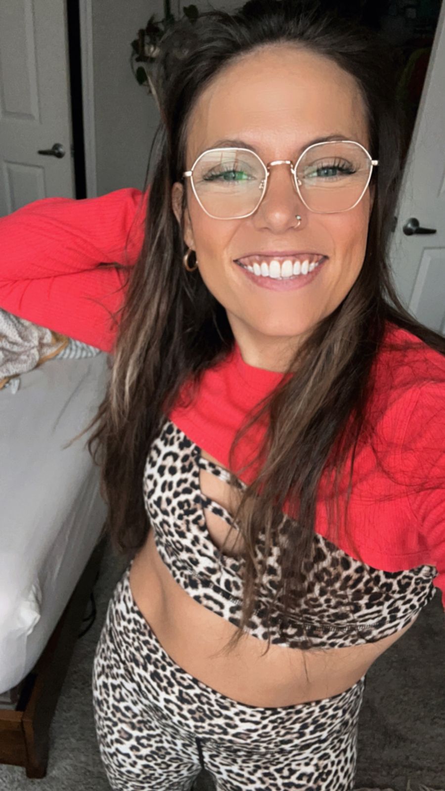 Woman with glasses and long brown hair wearing a red top and leopard print outfit, smiling at the camera in an indoor setting