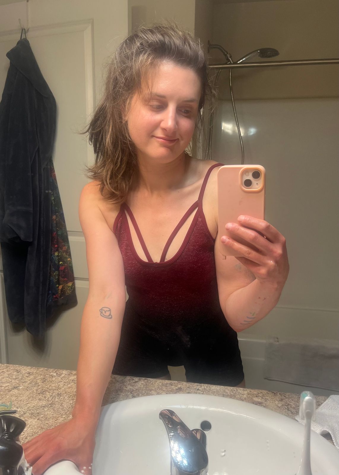 A person taking a mirror selfie in a bathroom, wearing a burgundy sleeveless top and holding a phone with a peach-colored case.