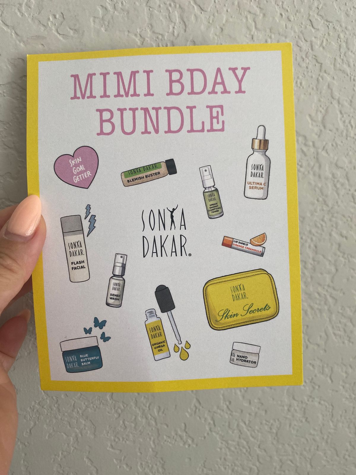 A hand holding a yellow-bordered card titled "MIMI BDAY BUNDLE" featuring illustrated Sonya Dakar skincare products including serums, cleansers, and other beauty items arranged around the brand logo