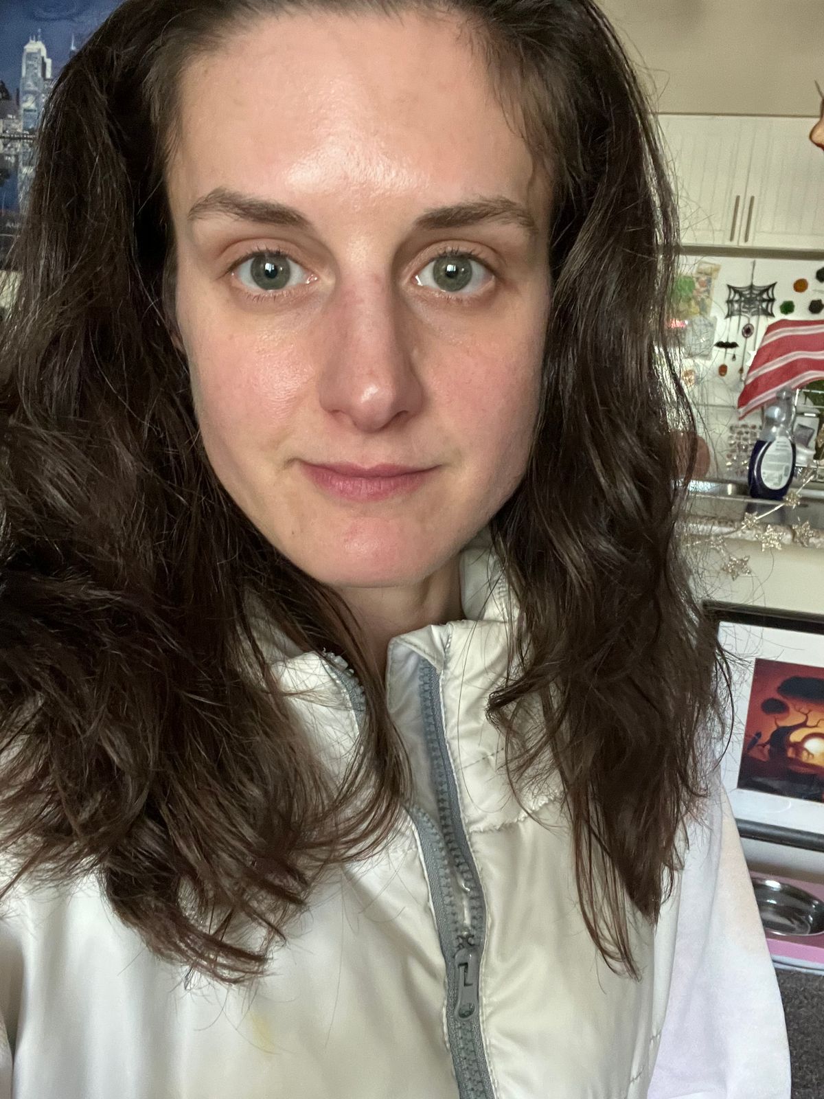 A person with long dark brown hair and green eyes, wearing a light-colored jacket with a zipper, taking a selfie indoors with shelving and decorative items visible in the background.