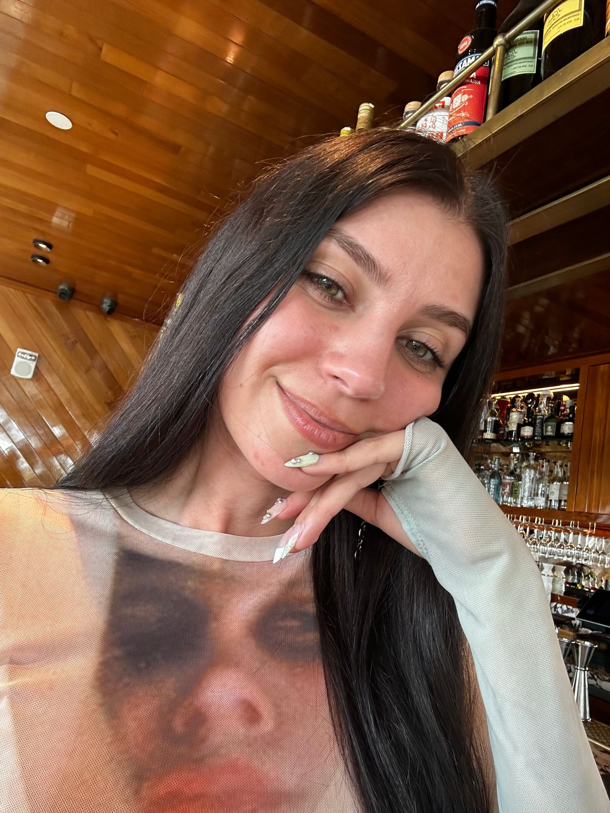 A woman with long dark hair smiling at the camera in what appears to be a restaurant or bar setting with wooden shelving and bottles visible in the background