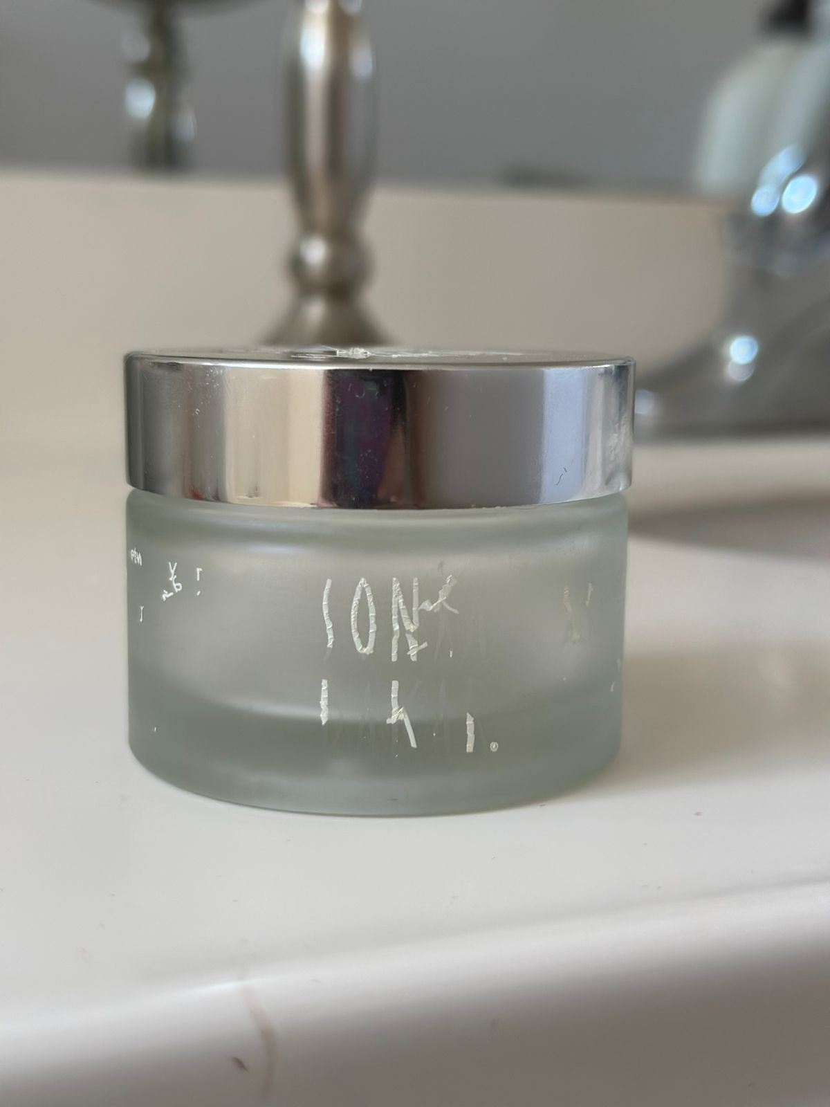 A frosted glass cosmetic jar with silver lid sitting on a white bathroom counter