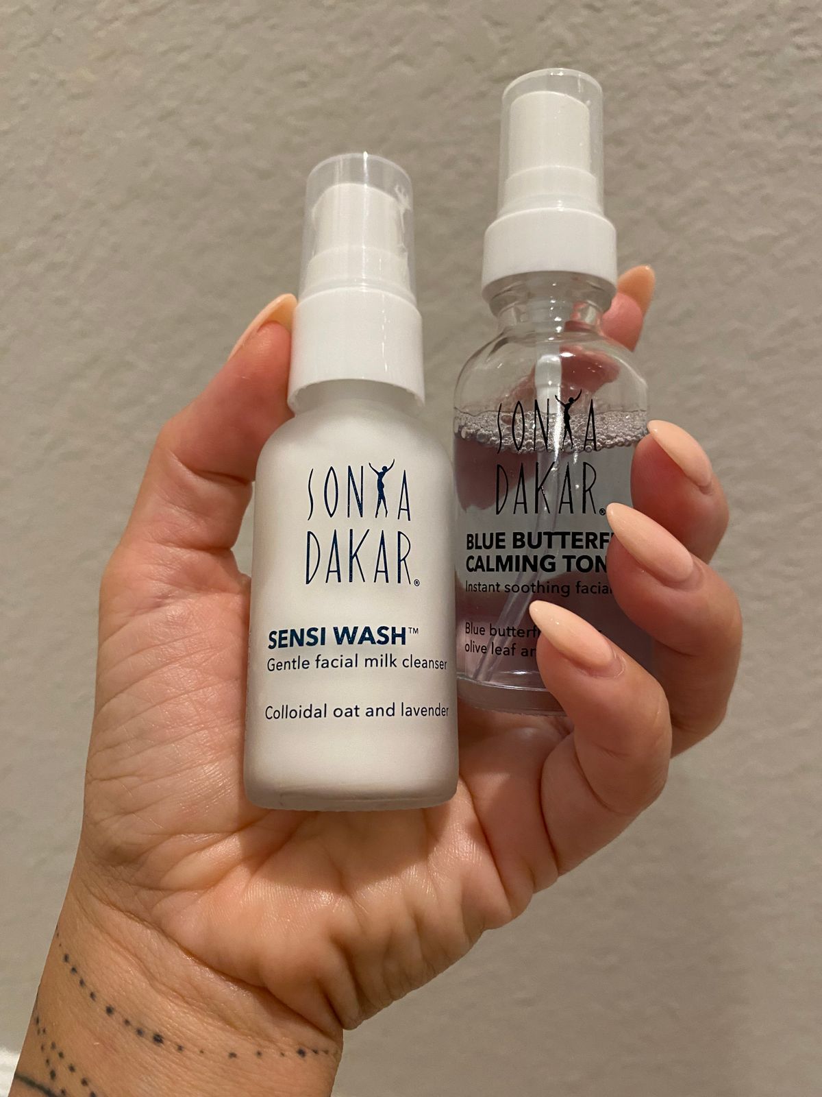 Two hands holding Sonya Dakar skincare products - a white bottle of Sensi Wash facial cleanser and a clear bottle of Blue Butterfly Calming Toner